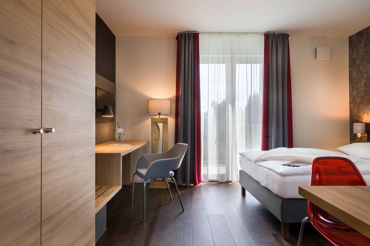 Een moderne hotelkamer met een houten garderobe, een bureau met stoel, een bed en een rode stoel.