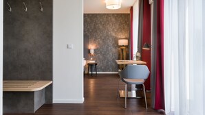 Een moderne hotelkamer met een houten bank, een zithoek met tafel en stoelen, een lamp en uitzicht buiten door dunne gordijnen.