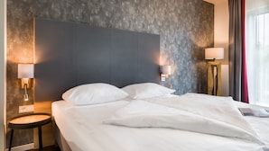 Een net opgemaakt bed met witte lakens in een moderne kamer, met een geruite grijze wand en een donker hoofdbord. Aan beide zijden van het bed staan twee bedlampjes.
