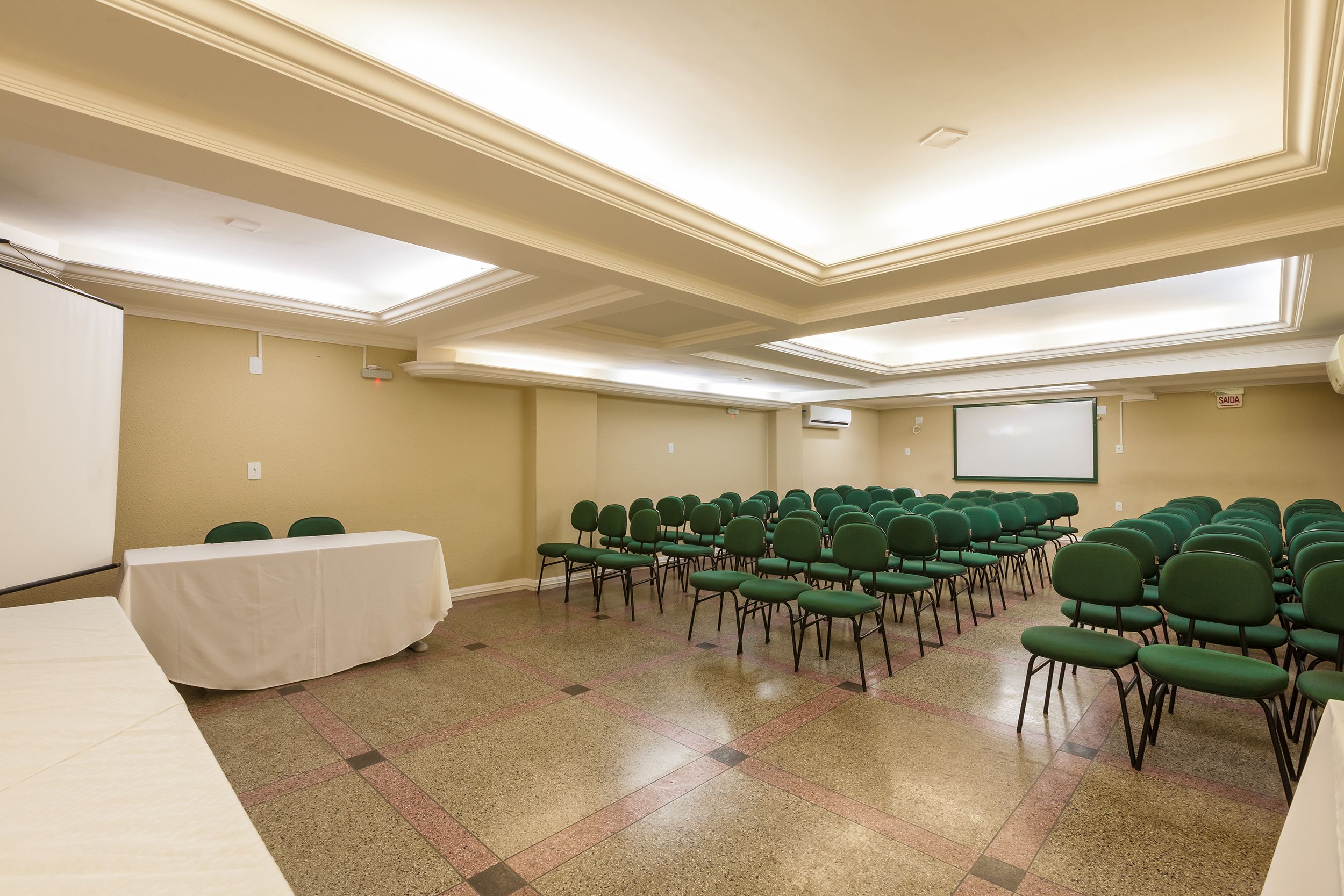 A conference room with a long table, rows of green chairs, a projector screen, and a whiteboard.
