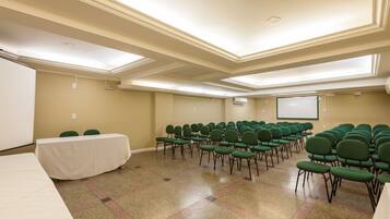 Un salón de conferencias con una larga mesa, filas de sillas verdes, una pantalla de proyector y una pizarra.