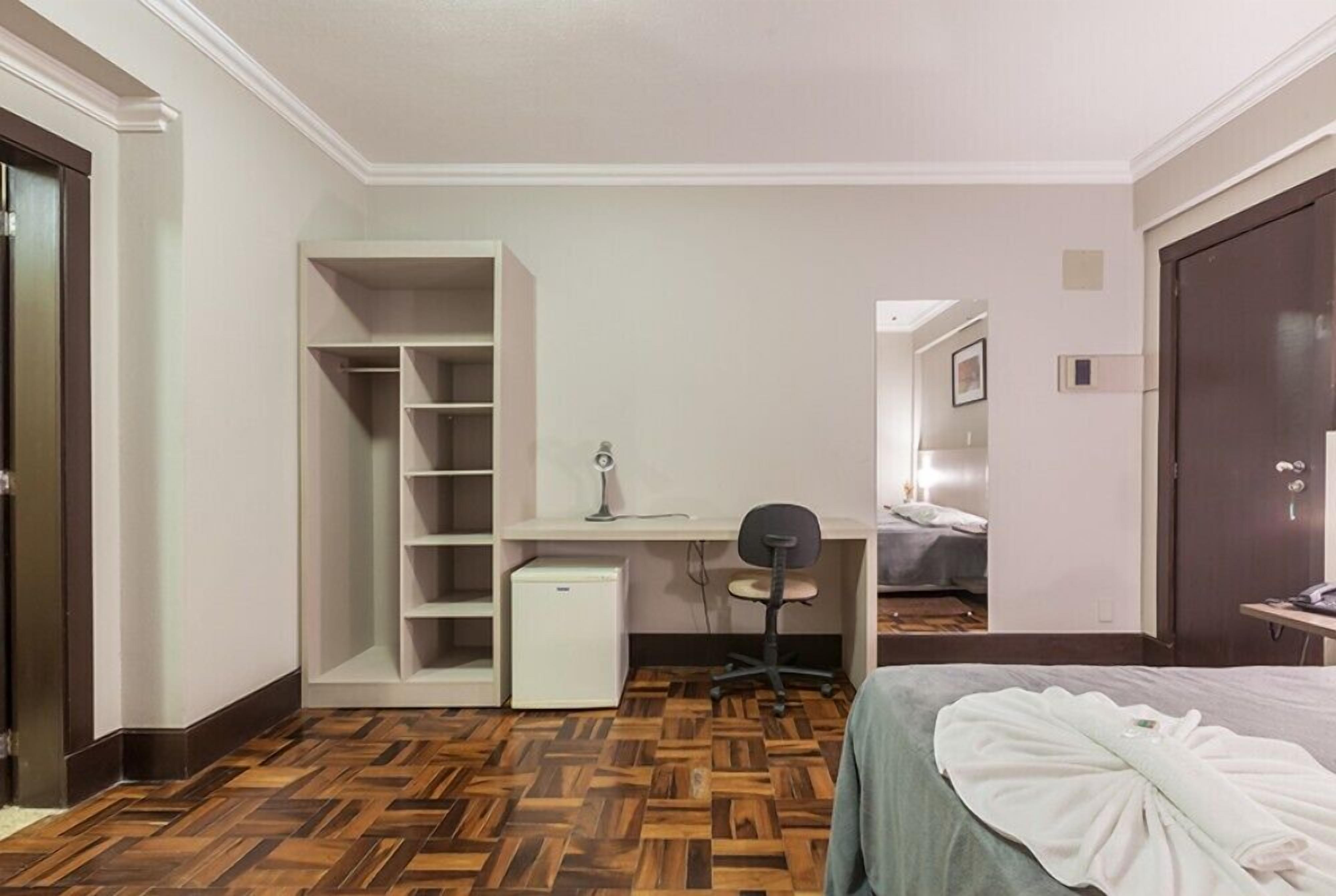 A hotel room with a wooden parquet floor, a desk with a chair, a wardrobe, and a bed with a white blanket.