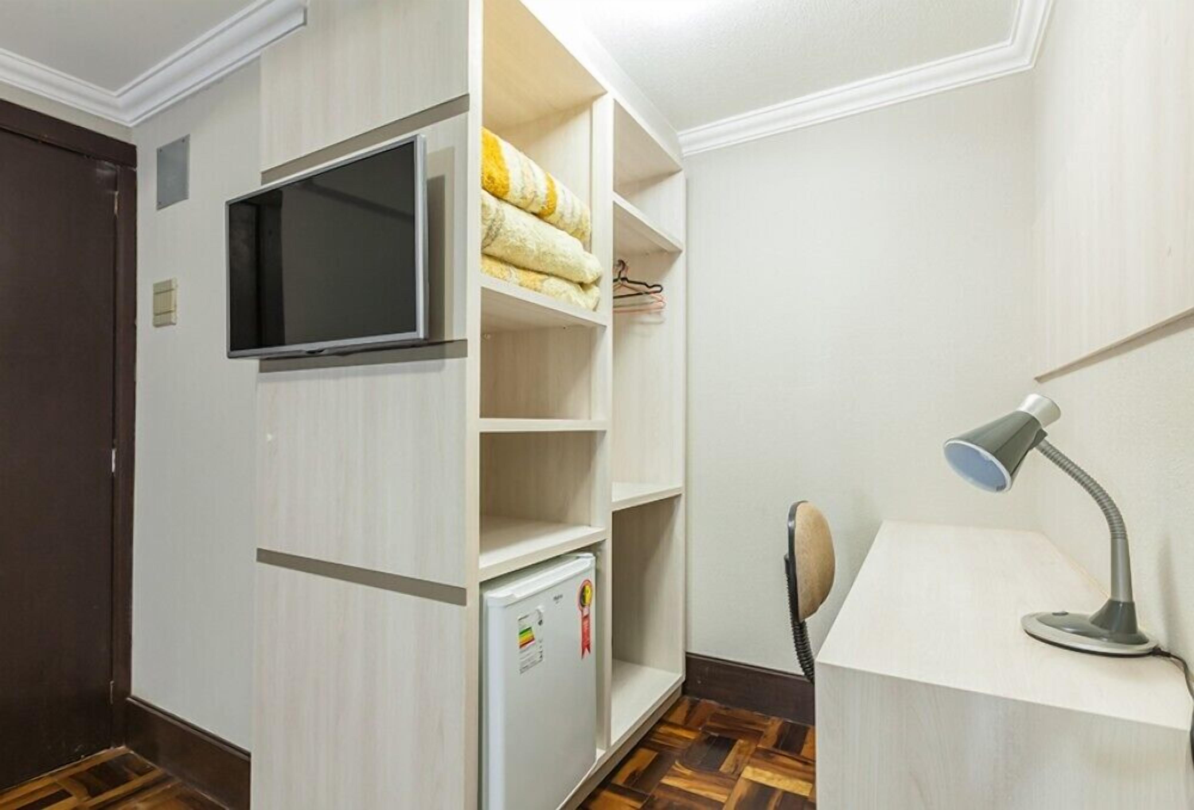 A compact room with a built-in TV, a small fridge, a desk with a lamp, and a shelving unit with towels.