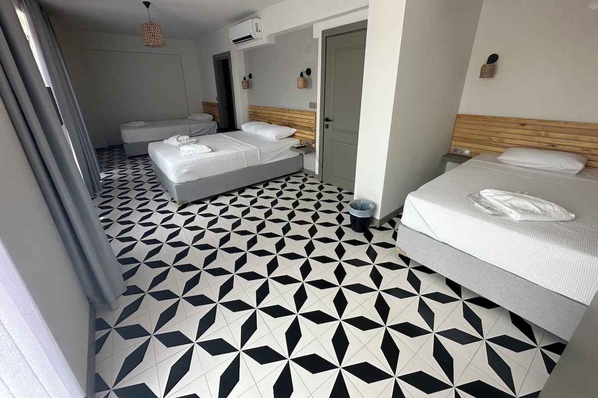 Habitación de hotel con piso de diseño geométrico, dos camas, cabecera de madera, unidad de aire acondicionado y un pequeño cesto de basura.