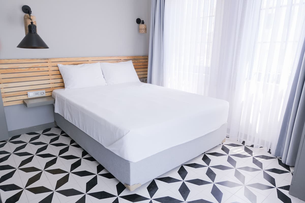 Una cama bien hecha en una habitación con suelo de estampado geométrico, un cabecero de madera y una lámpara fijada a la pared.