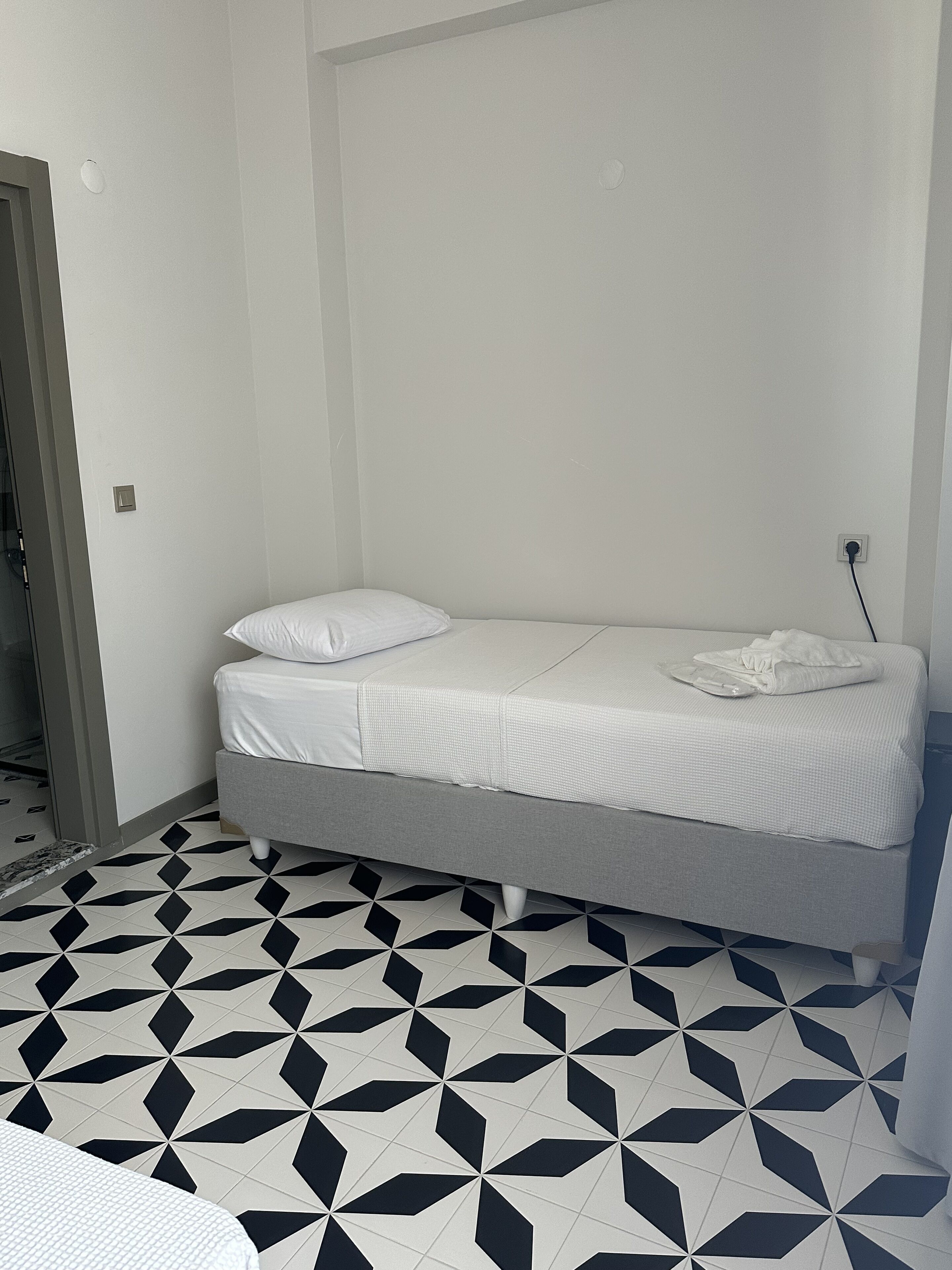 A small, single-bed room with a geometric patterned floor, a grey bed, and a white wall.