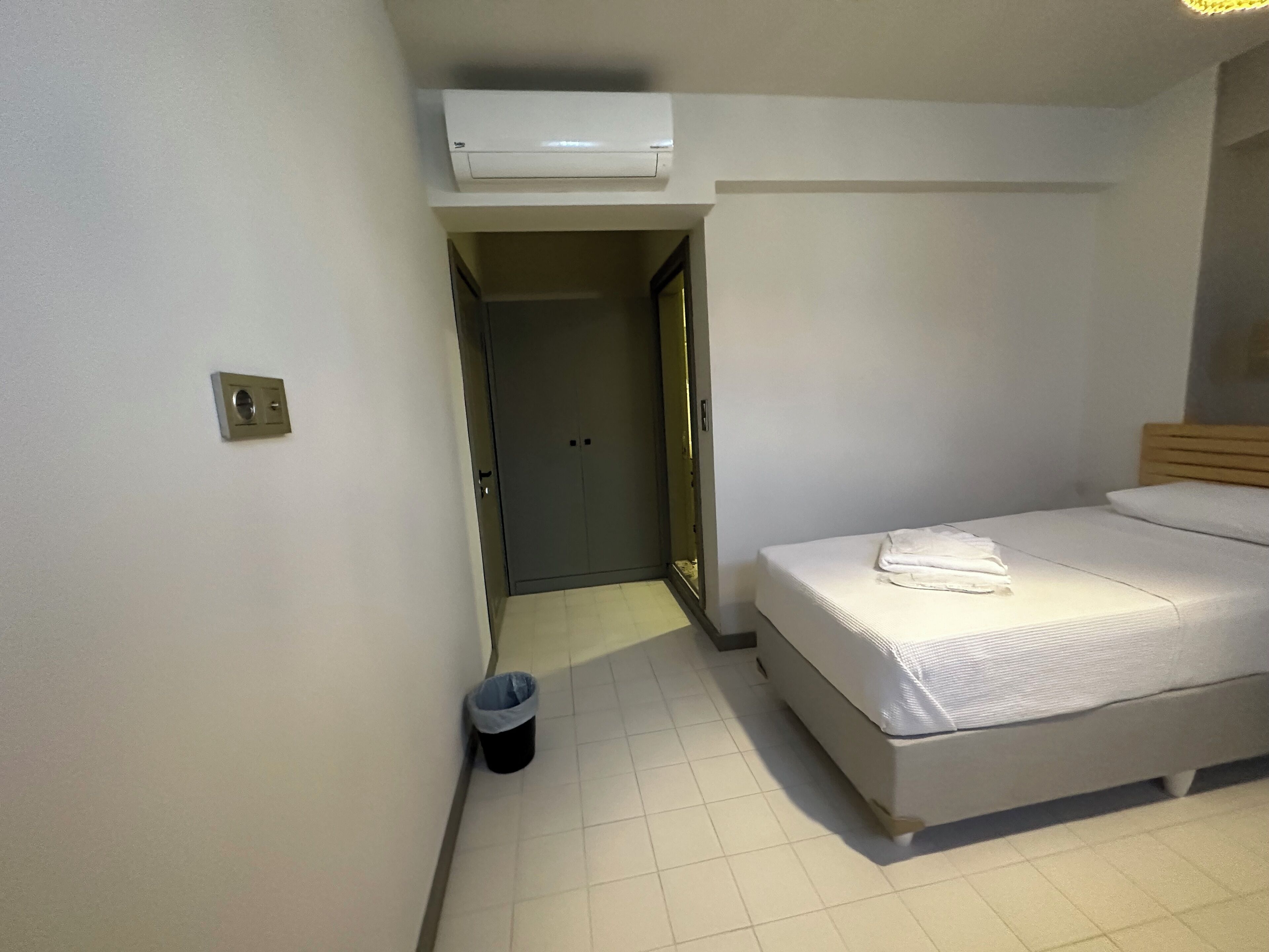 A hotel room with a bed, a trash bin, an air conditioning unit, and a door leading to another room.