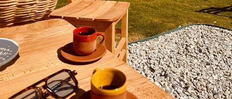An outdoor seating area with wooden tables and chairs, a wicker chair, and a small table with a cup and saucer.