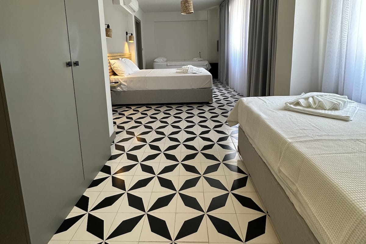 Habitación de hotel con suelo de estampado geométrico, cama con edredón blanco, armario y lámpara colgante.