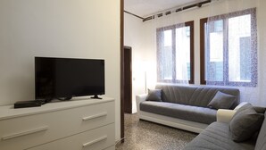 A modern living room with a flat-screen TV on a white cabinet, a grey sofa, and a patterned window treatment. - Coco (Venice)