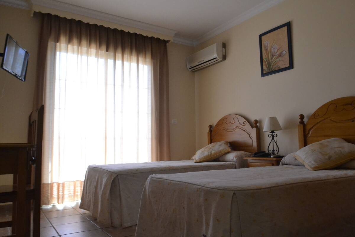 Une chambre d’hôtel avec deux lits simples, une tête de lit en bois, une table de chevet avec une lampe, un téléviseur fixé au mur et un appareil de climatisation.