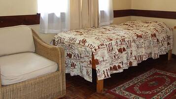 Een kamer met een enkel bed, een rieten fauteuil, een klein houten tafeltje en een kamerbreed tapijt met dessin.