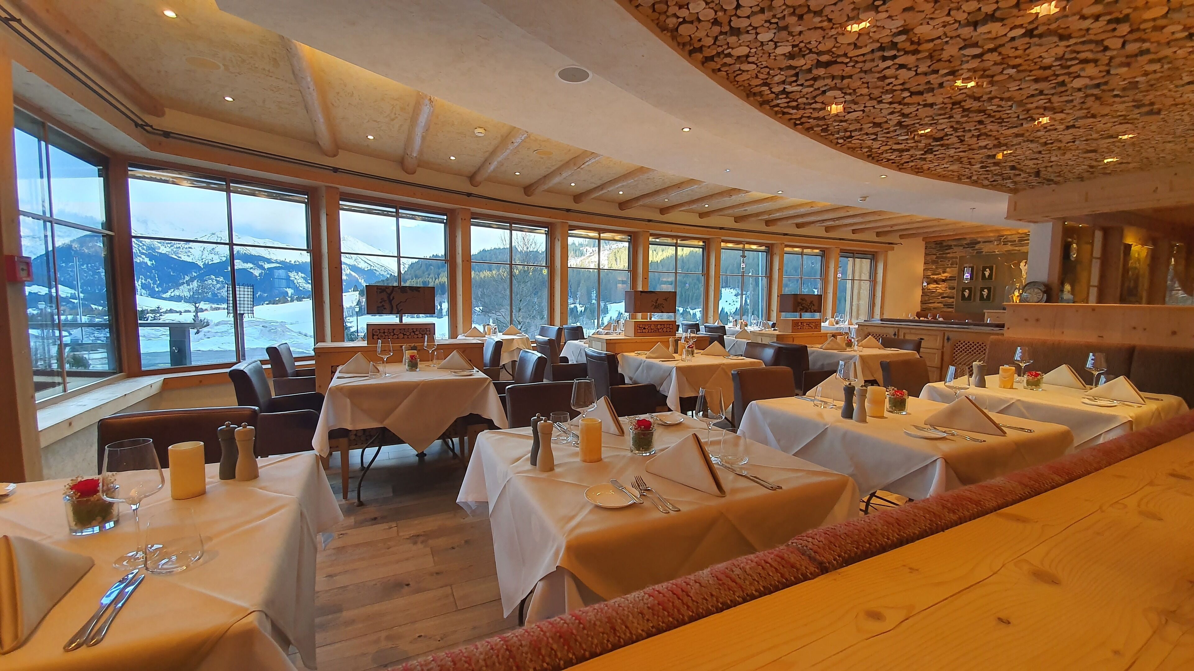 Ein Restaurant mit mehreren Tischen, großen Fenstern mit Blick auf die Berge und einer Holzbalkendecke.