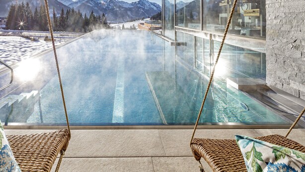 Une piscine offrant une vue dégagée sur les montagnes enneigées, un espace clos par des parois de verre et des fauteuils en osier dotés de coussins.