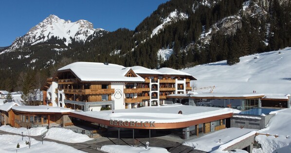 Un hôtel à plusieurs étages, avec un télésiège visible en arrière-plan, entouré de montagnes enneigées.