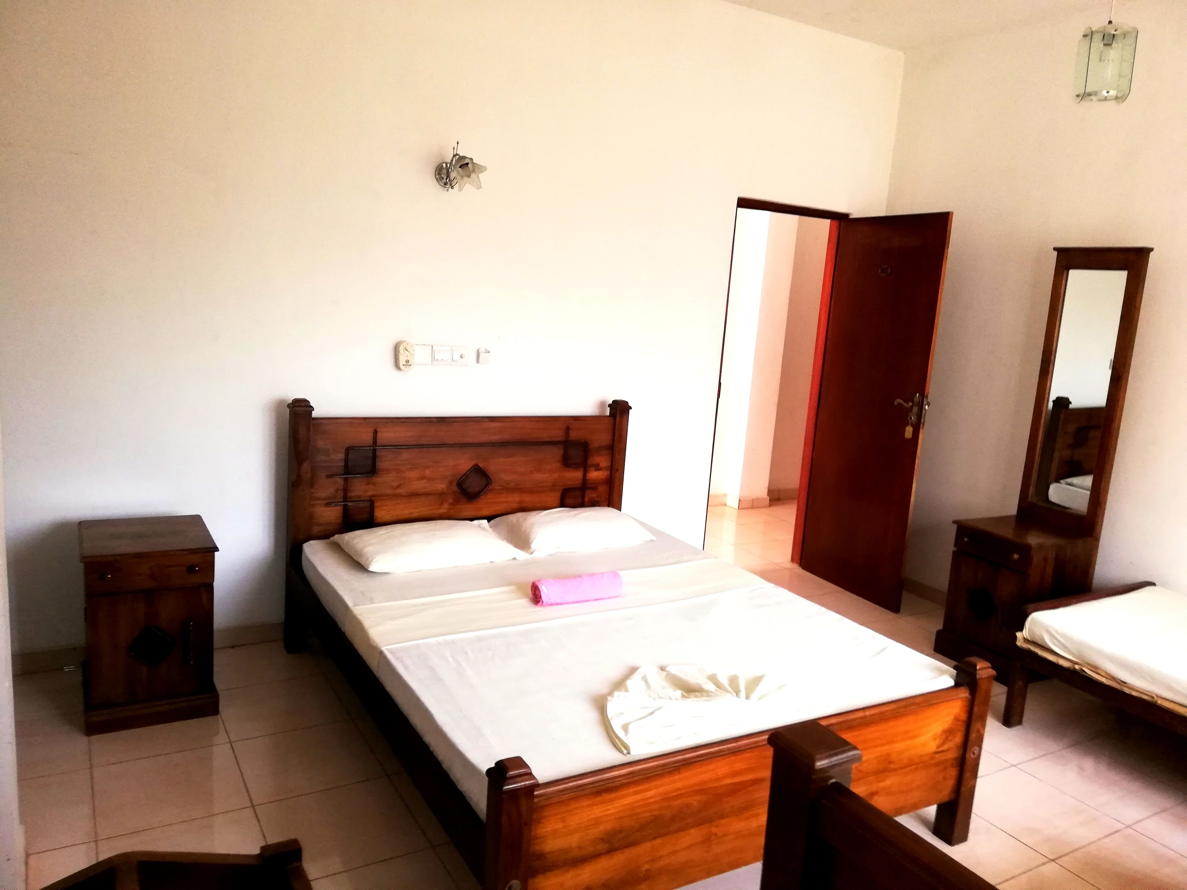 A bedroom with a wooden bed, a bedside table, a nightstand, a mirror, and a door leading to another room.