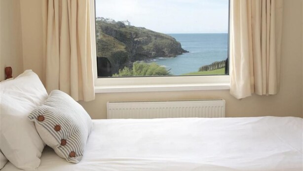 A bedroom with a bed, two pillows, a window with a view of a coastal landscape. - Green Door - Lower Tregudda (Port Isaac)
