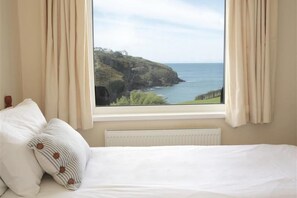 A bedroom with a bed, two pillows, a window with a view of a coastal landscape. - Green Door - Lower Tregudda (Port Isaac)