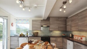 A modern kitchen with wooden cabinets, a dining table with chairs, and a window with a view of greenery. - Green Door - Lower Tregudda (Port Isaac)