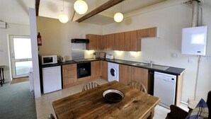 Una cocina compacta con armarios de madera, microondas, lavaplatos y un fregadero. Hay una mesa de madera con un bol encima, y se ve una silla.