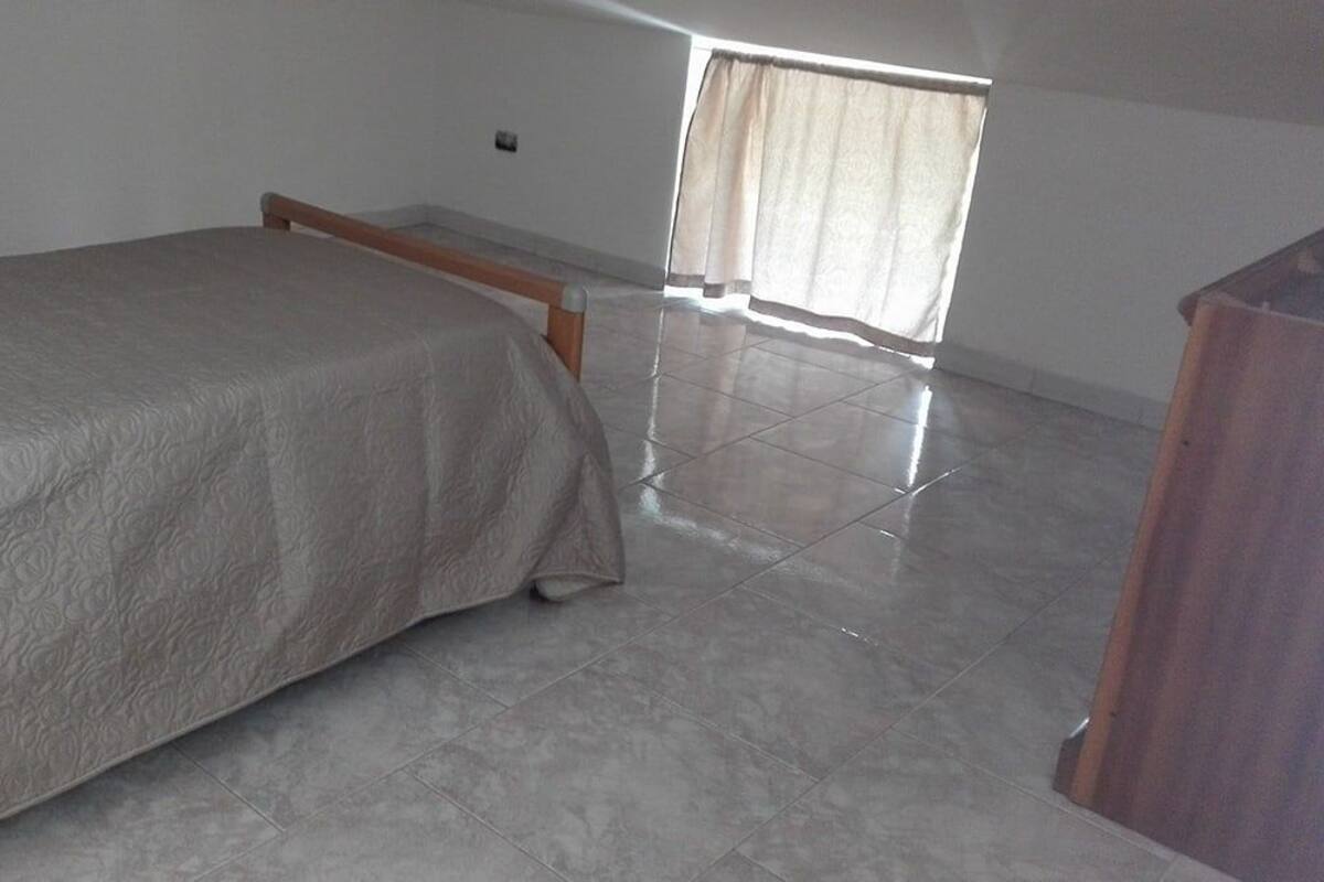 A bedroom with a bed, a wooden headboard, a window with curtains, and a tiled floor.