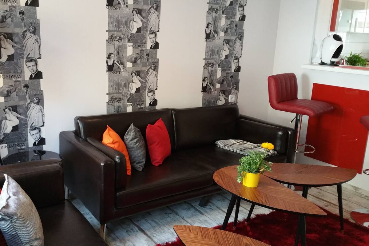 A living room with a dark brown leather sofa, red and grey cushions, a round wooden coffee table with a yellow plant, and a red bar stool.