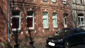 Un edificio de varios pisos de ladrillo rojo, con varias ventanas, un coche estacionado adelante y un cielo azul despejado.