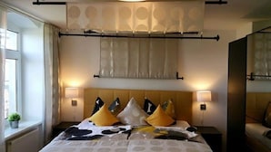 Habitación de hotel con cama, dos lámparas de noche, ventana con cortinas y cabecero con almohadones decorativos.