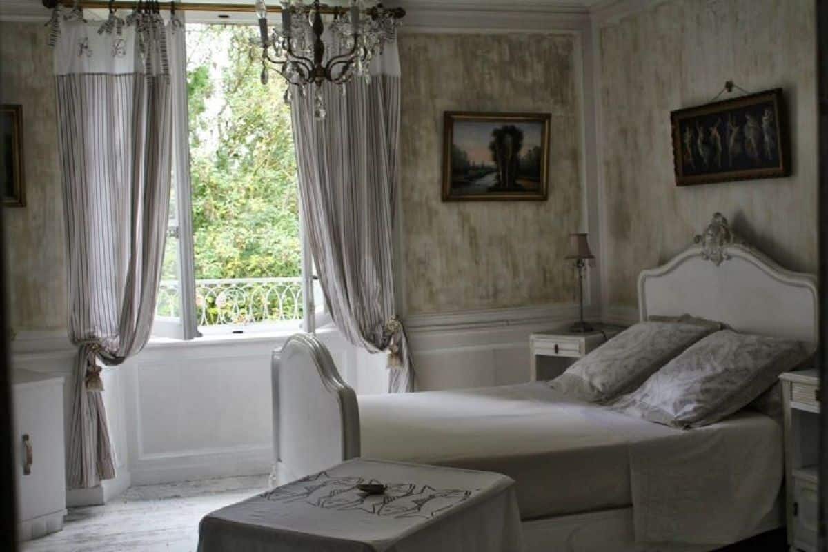A bedroom with a large bed, a bedside table, a chair, and curtains. There is a window with a view of greenery outside.