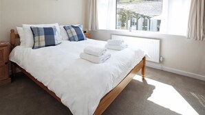 A neatly made bed with white linens and folded towels, a wooden headboard, and a view of a green landscape through a window. - Green Door - Marigold (Port Isaac)