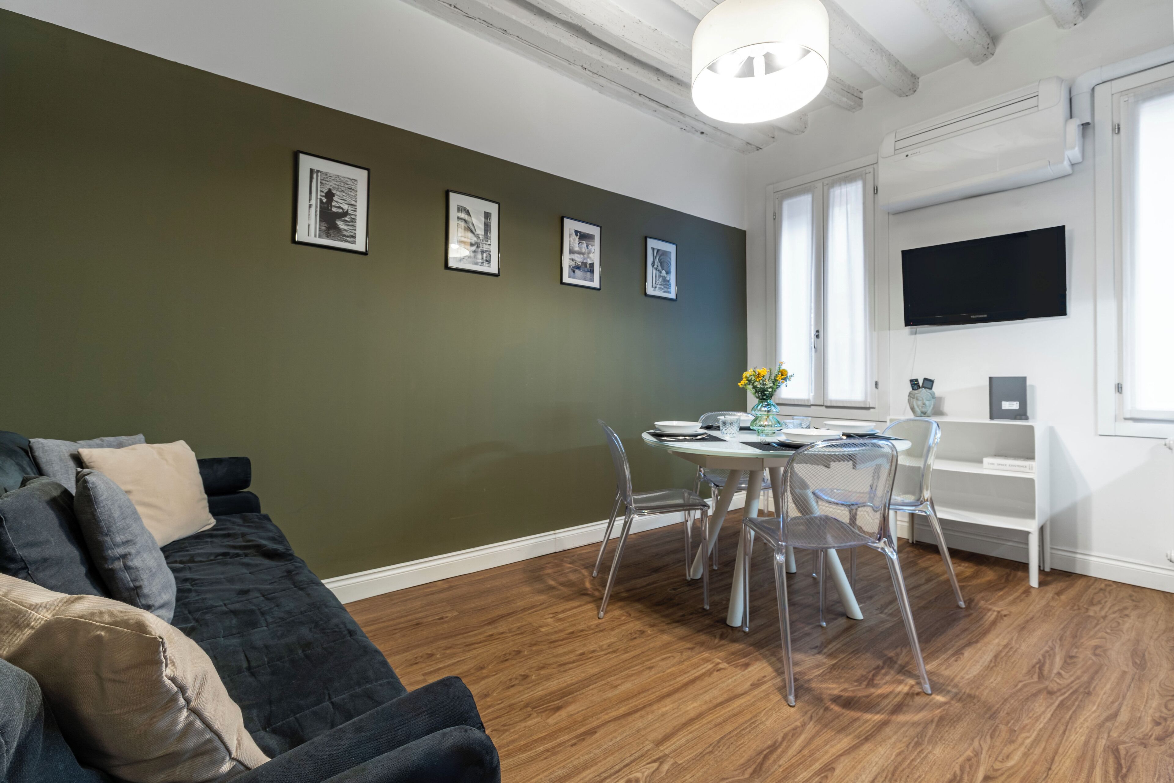 Una sala de estar con una pared verde oscuro, un sofá negro con cojines beige, una mesa de comedor con sillas transparentes y un televisor de pantalla plana montado en la pared.
