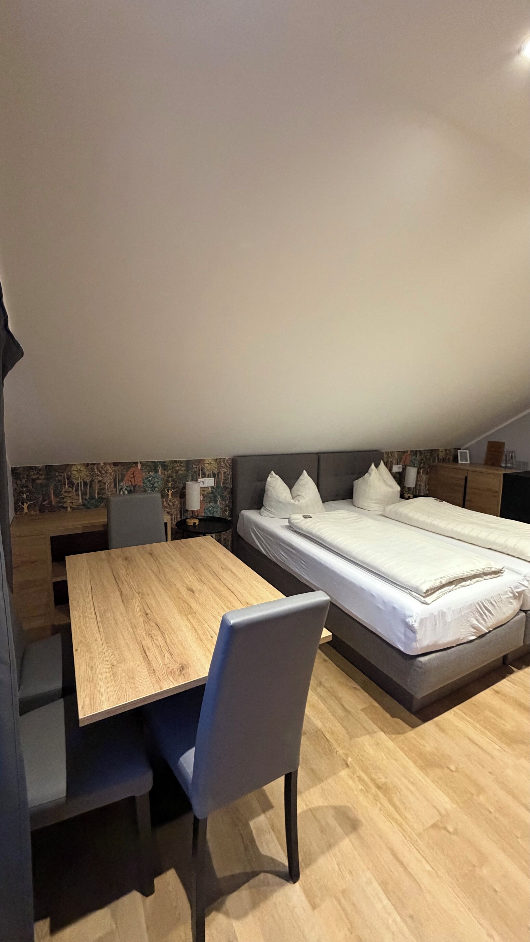 Un dormitorio moderno con una mesa de comedor de madera, una única silla gris, una cama grande con ropa de cama blanca y un mural en la pared.