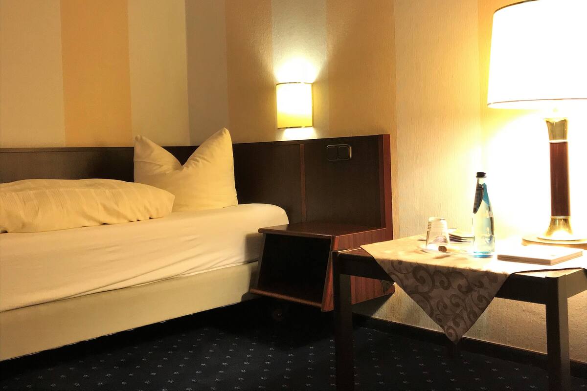 A hotel room with a bed, a nightstand, a table with a lamp, and a wall-mounted light.