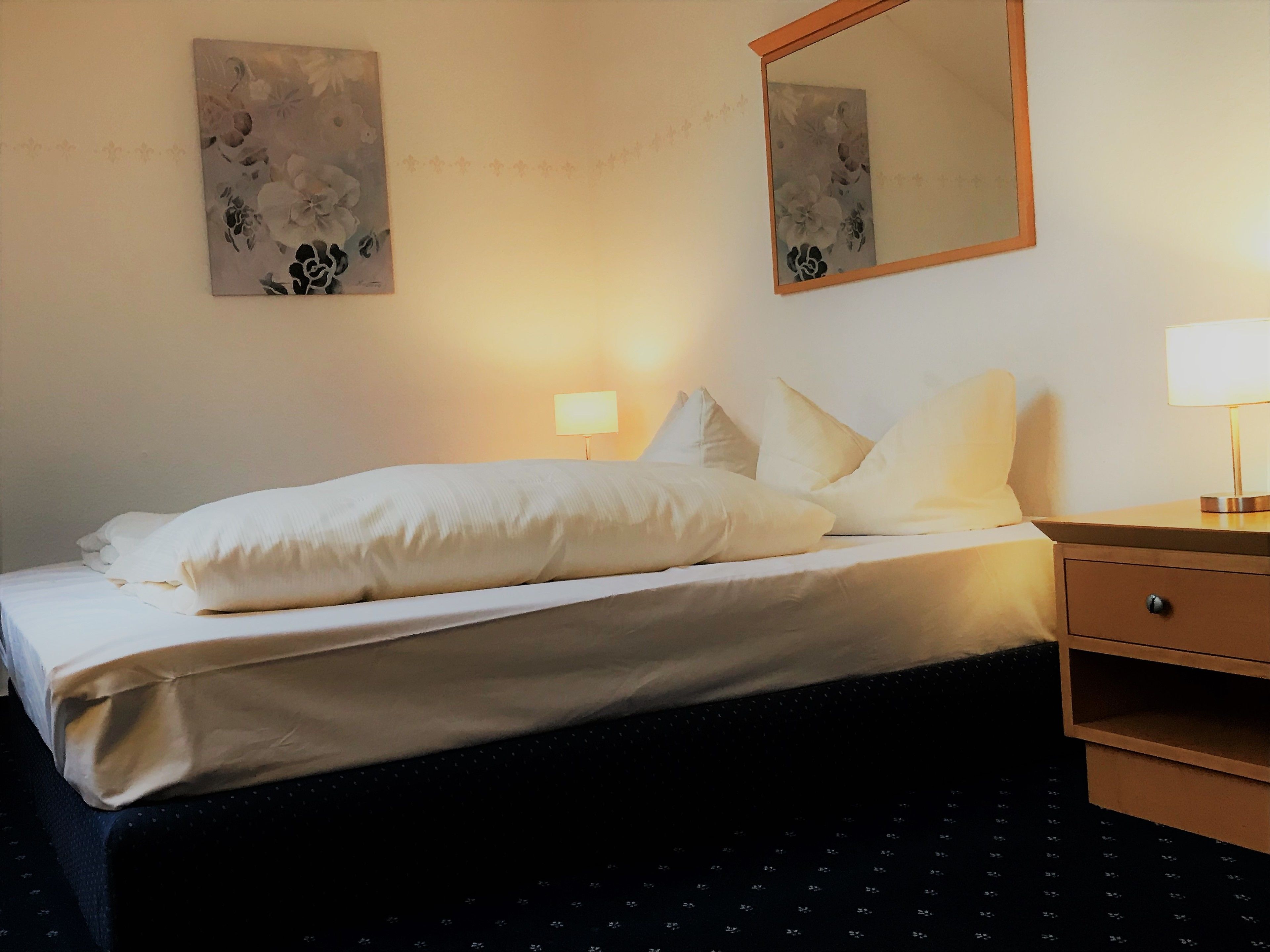 Una habitación de hotel con una cama, una mesita de noche, una lámpara y dos cuadros colgados en la pared.