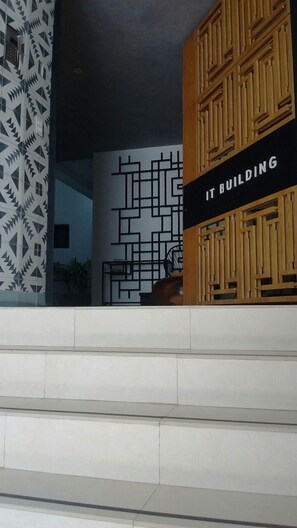 Fachada moderna de un edificio con un muro de diseño geométrico, un panel de madera con la señalización 'IT BUILDING' y una escalera que sube.