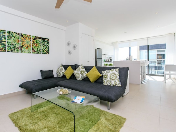 A modern living room with a dark grey sofa, green and yellow patterned cushions, a glass coffee table, and a green rug.