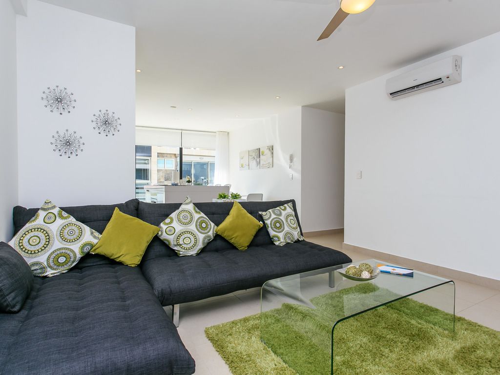 A modern living room with a grey sofa, green and patterned cushions, a glass coffee table, and a green rug.