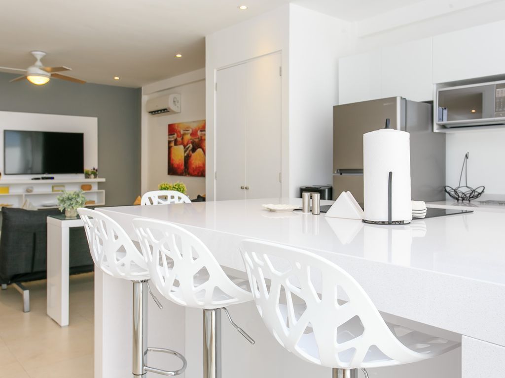 A modern kitchen with white cabinetry, a central island with bar stools, a flat-screen TV, and a ceiling fan.