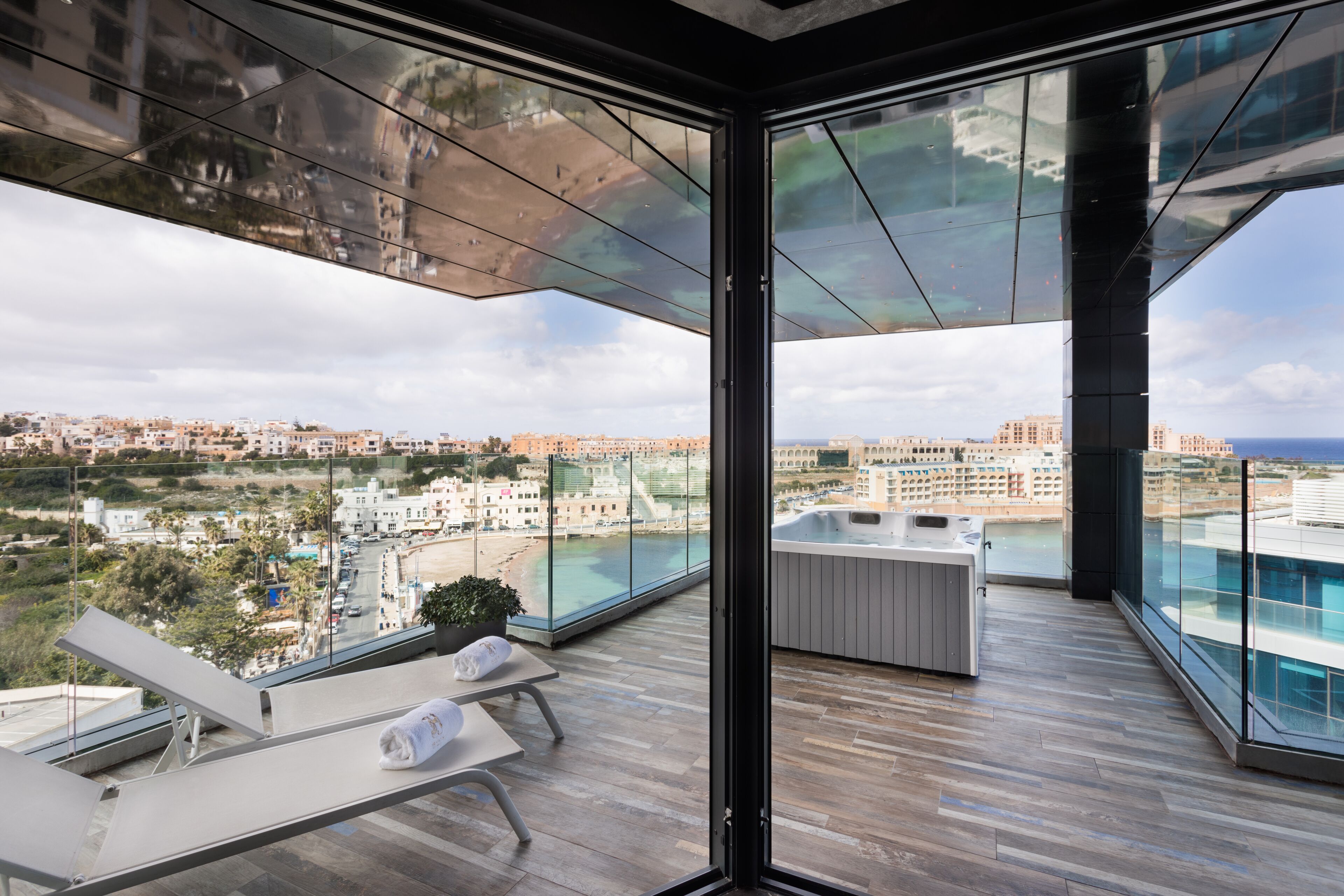 A modern balcony with a glass enclosure, offering a view of a coastal cityscape and a swimming pool.
