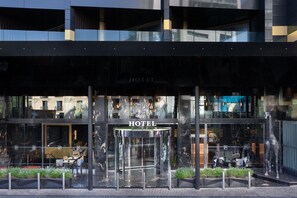 A modern hotel entrance with a glass facade, a revolving door, and outdoor seating. - H Hotel (St. Julian's)