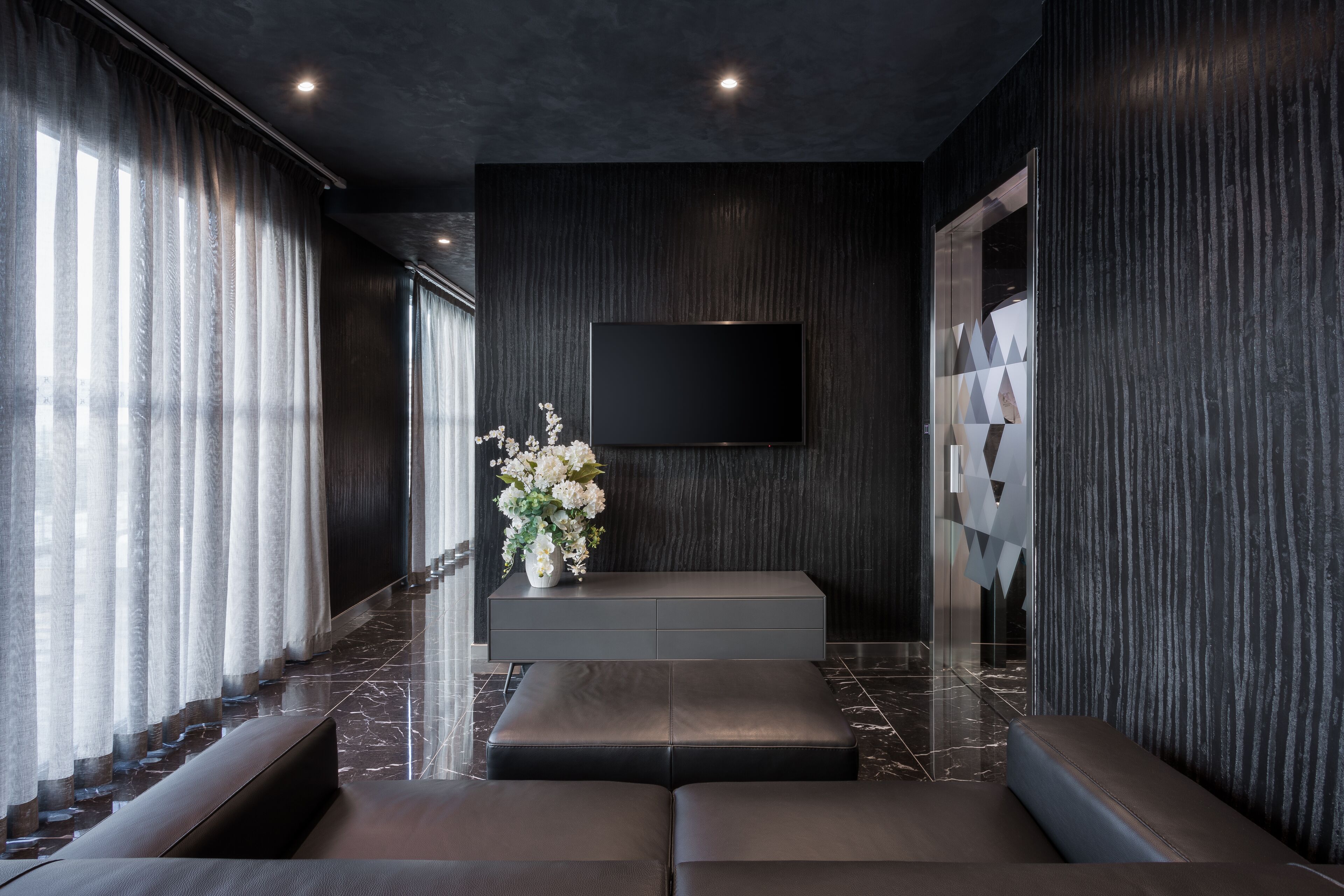 Una hall moderna con parete rivestita in legno scuro, una TV a schermo piatto montata sopra una consolle grigia, pavimento in marmo e una zona relax con un vaso di fiori bianchi.