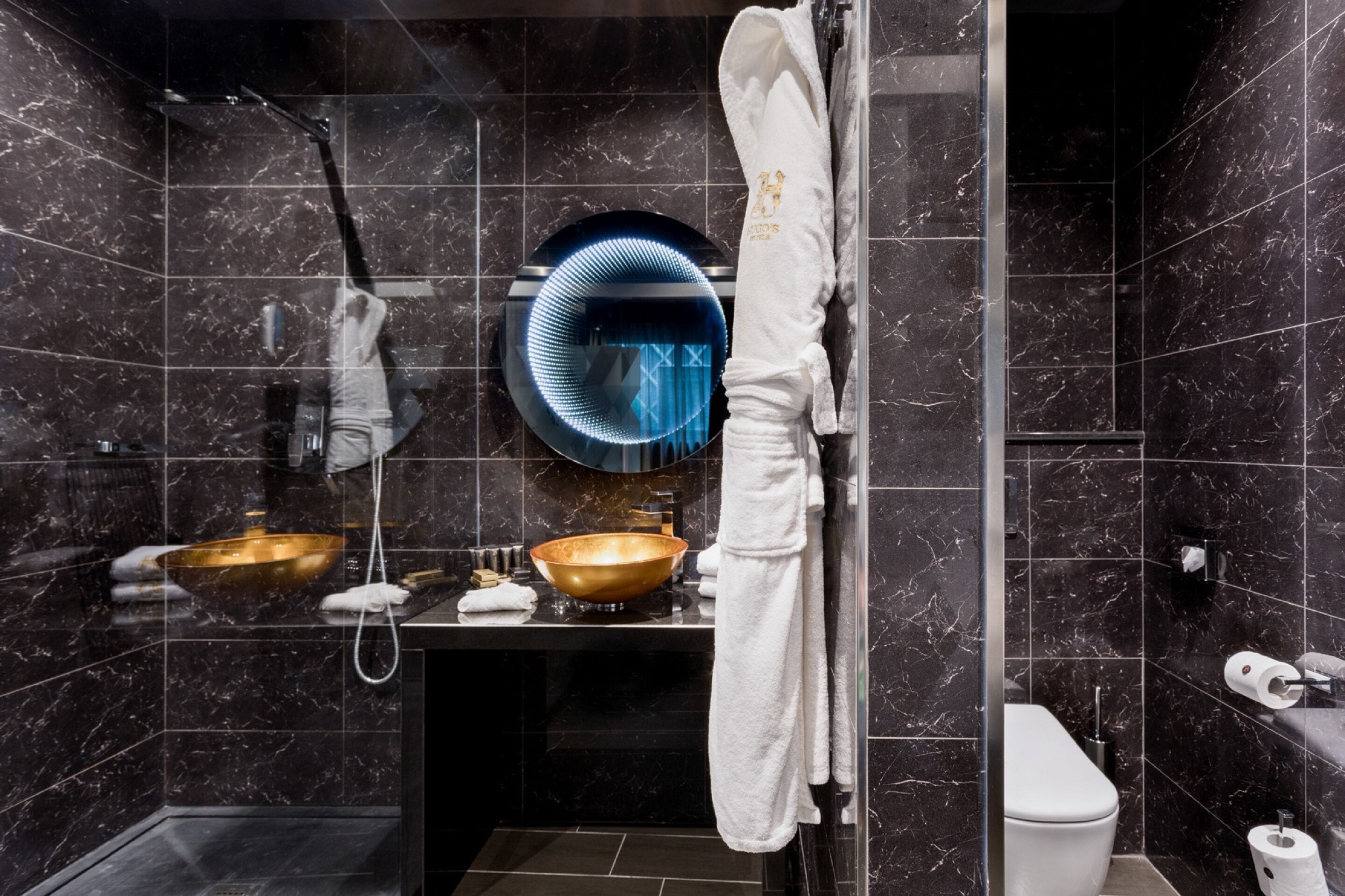 A modern bathroom with dark marble tiles, a gold basin, a round mirror, and a white towel hanging on a rack.