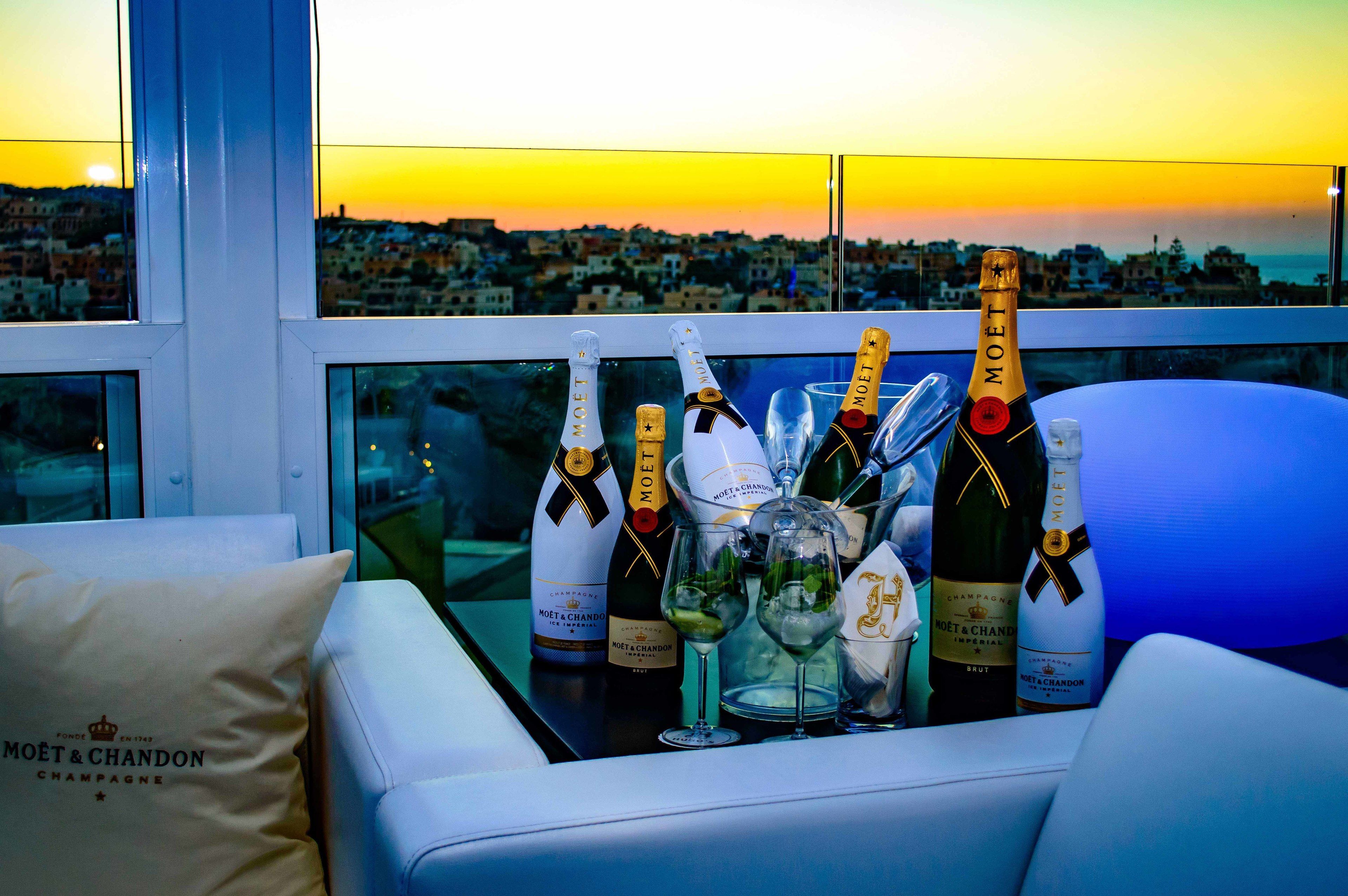 A table with multiple bottles of Moët & Chandon champagne, glasses, and a bottle of Brut champagne, set against a cityscape view at sunset.