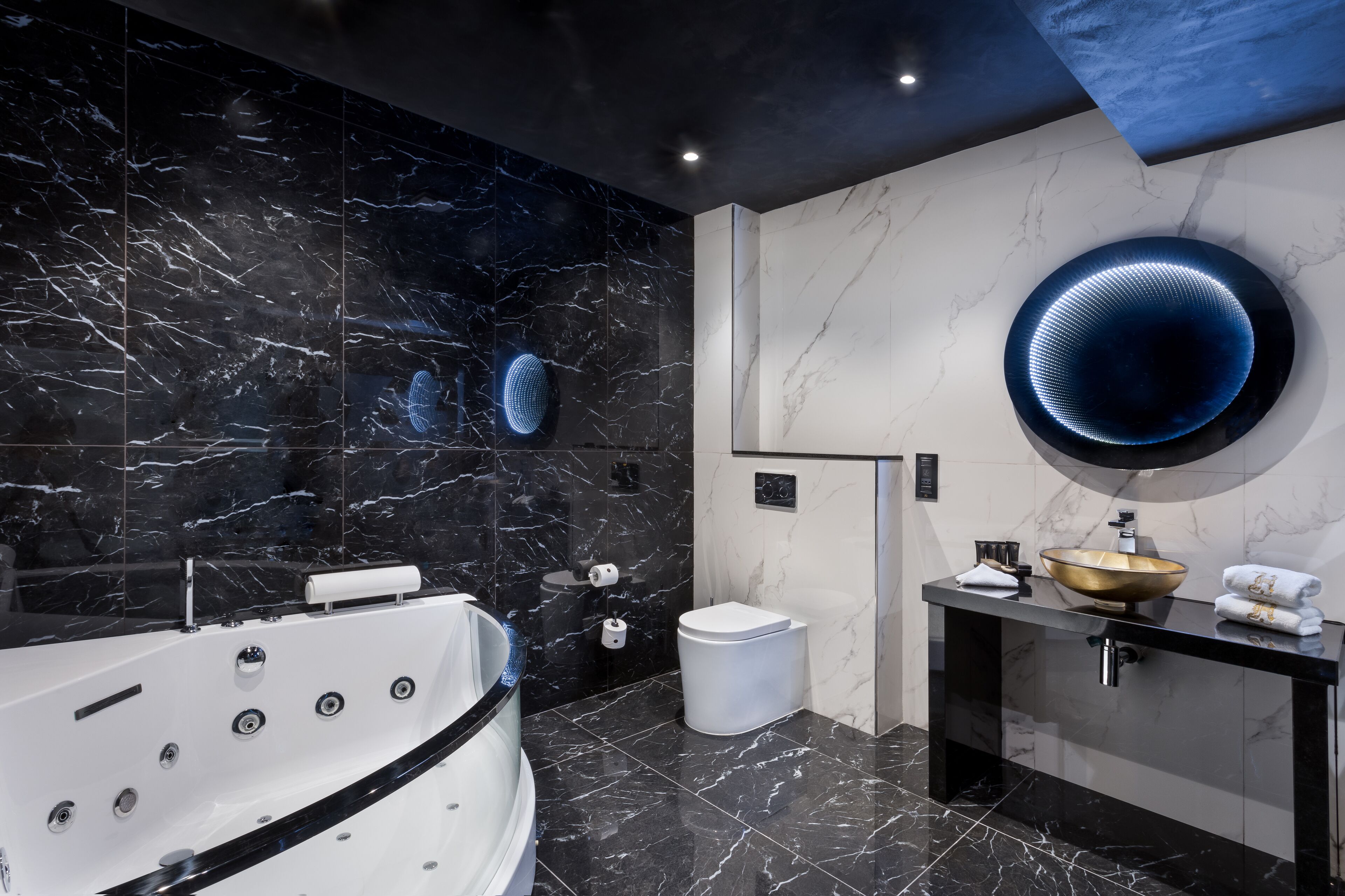 A modern bathroom with a black and white marble design, a large round mirror, a freestanding bathtub, and a sleek vanity with a gold bowl sink.