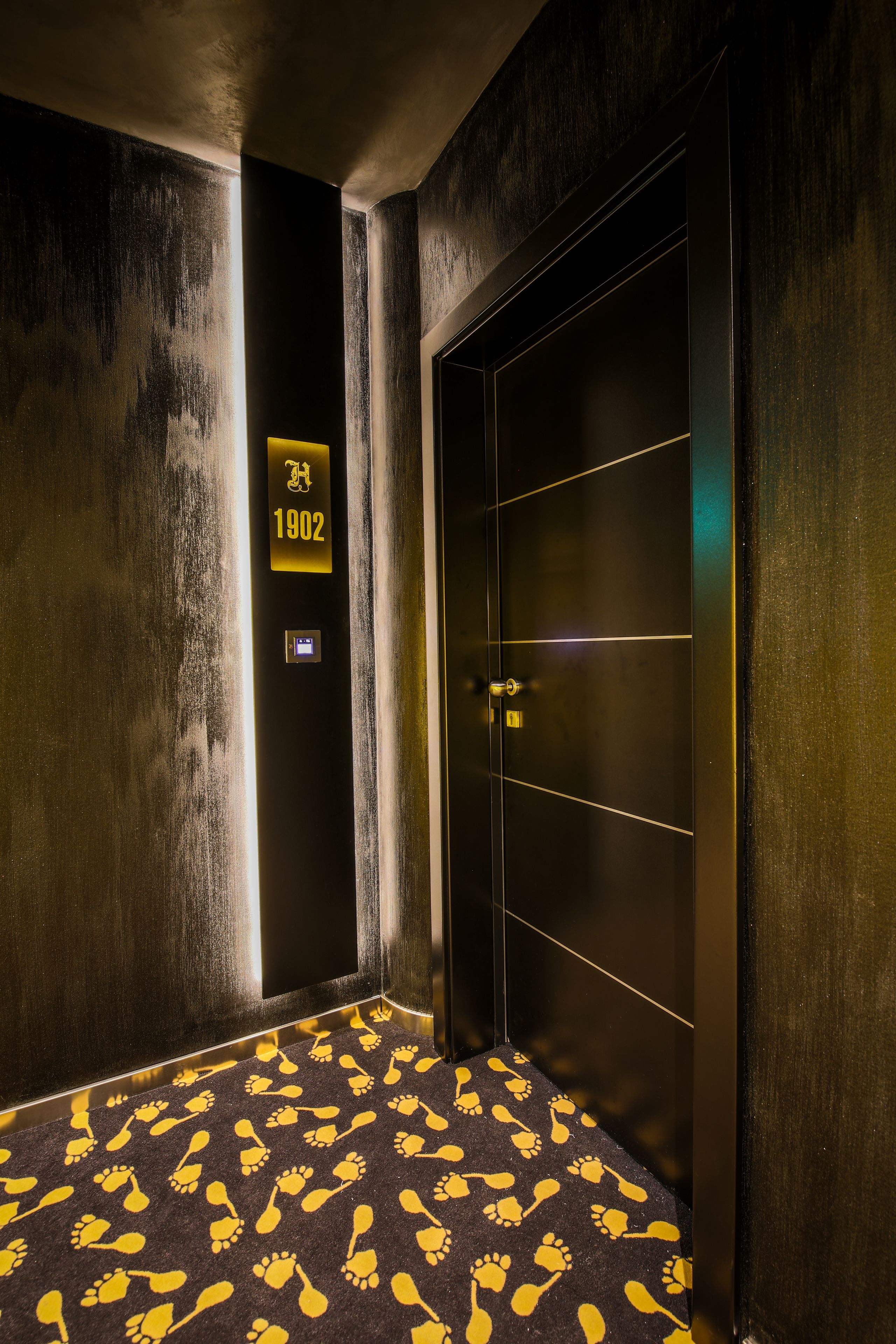 Un pasillo con piso oscuro, una puerta con el letrero "1902" y una pared con textura.
