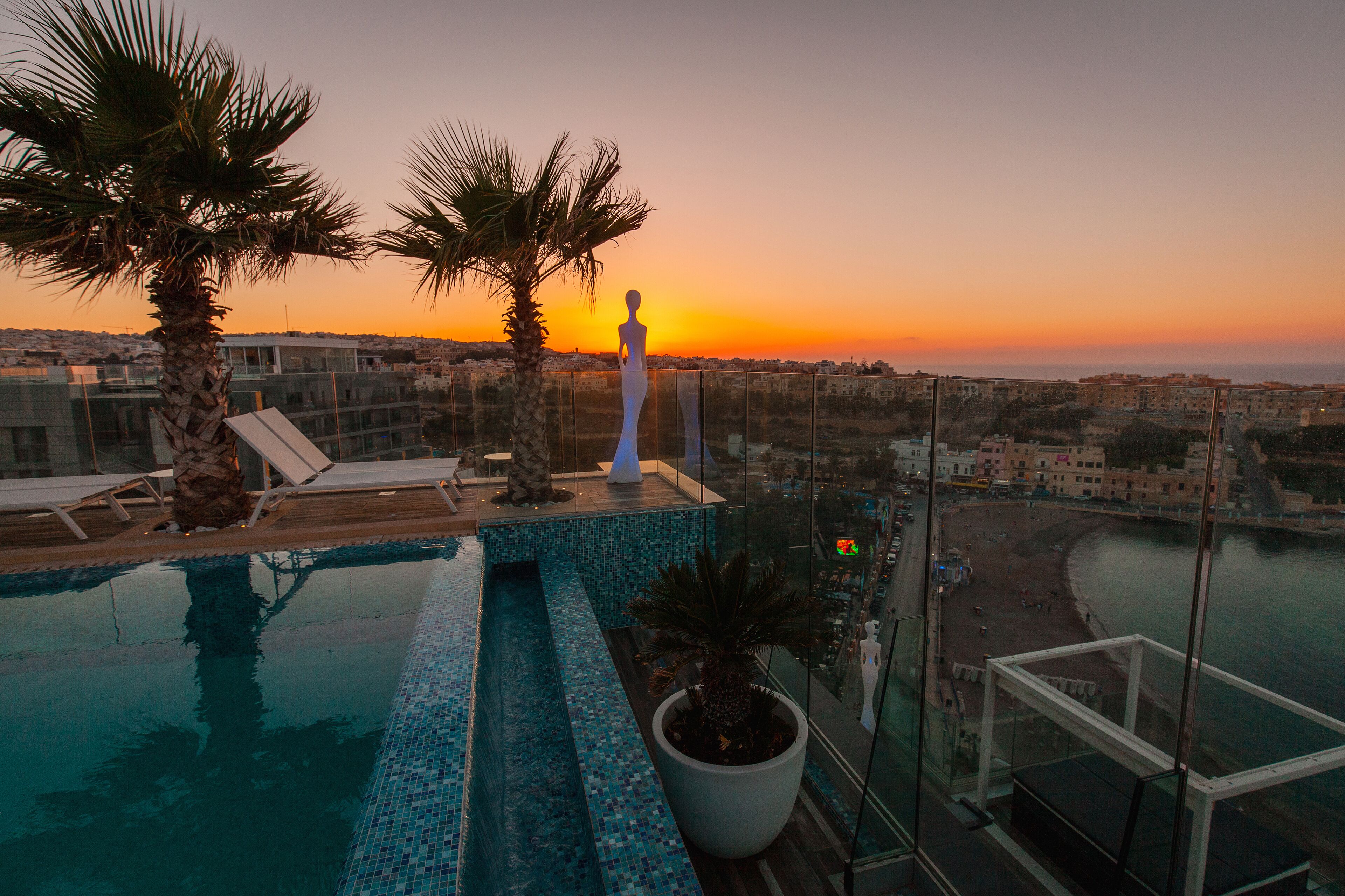 A rooftop infinity pool with a view of the sunset over a coastal cityscape.