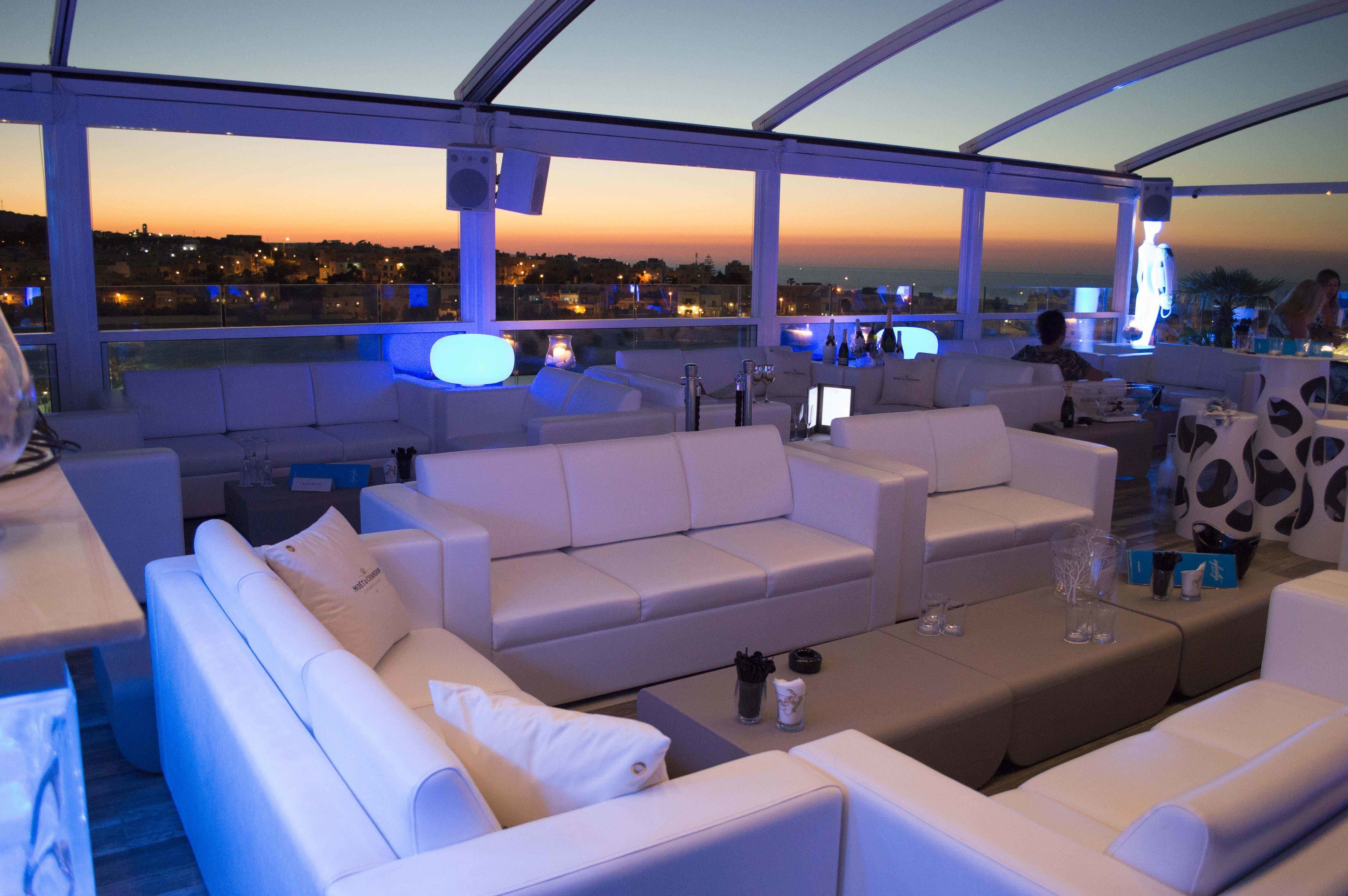 A rooftop lounge with white sofas and tables, a glass balustrade, and a city view at dusk.