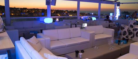 A rooftop lounge with white sofas and tables, a glass balustrade, and a city view at dusk.