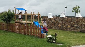 Un parque infantil con una estructura de madera para escalar y un tobogán azul, rodeado por un muro de piedra y una valla de madera.