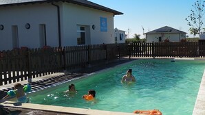 Zona de piscina con valla de madera, edificio con un letrero y área verde con una mesa de picnic.